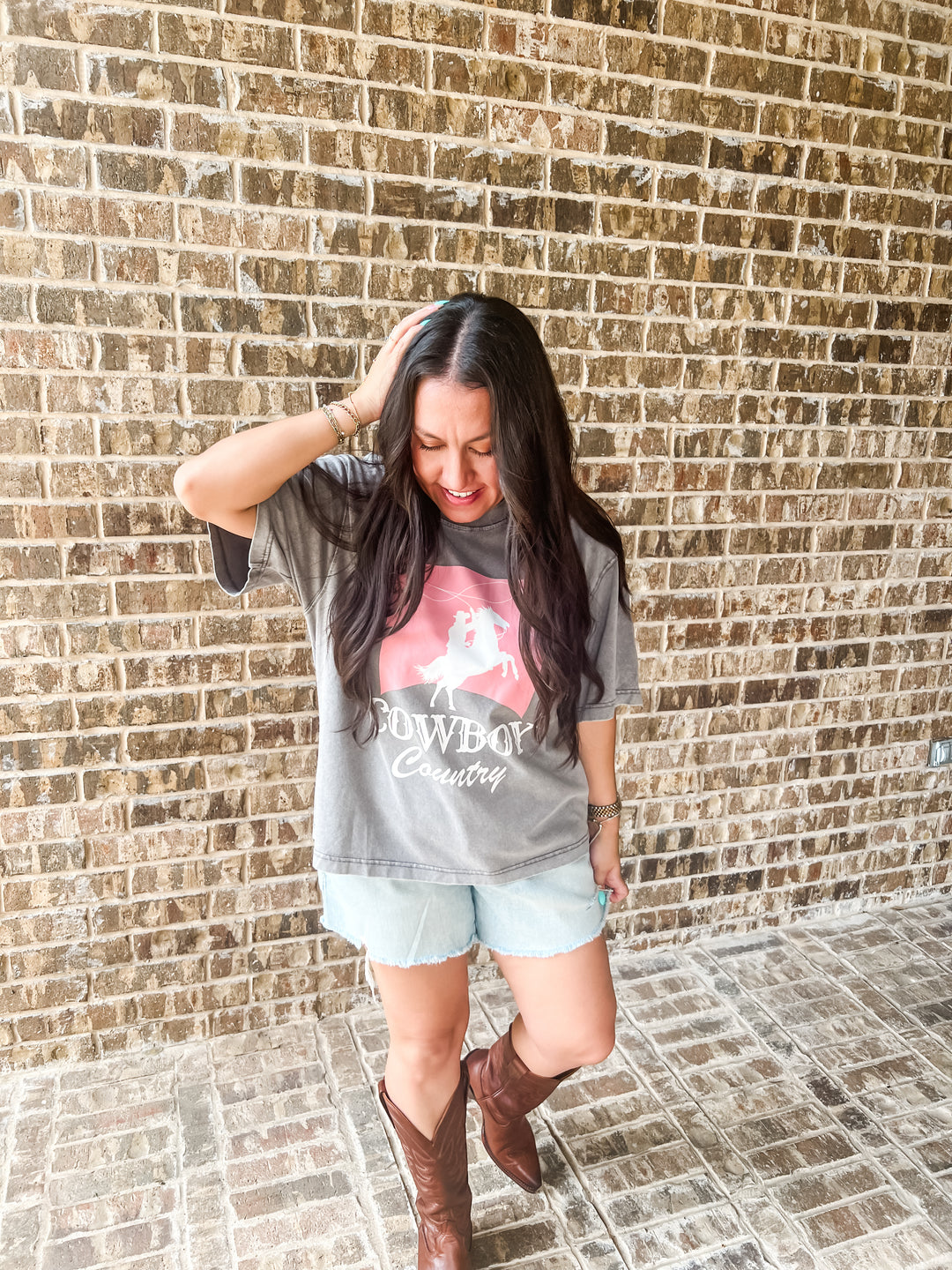 A woman posing in front of a brick wall wearing a gray graphic t-shirt with a pink and white cowboy design that says "COWBOY COUNTRY", light blue denim shorts, and brown leather cowboy boots.