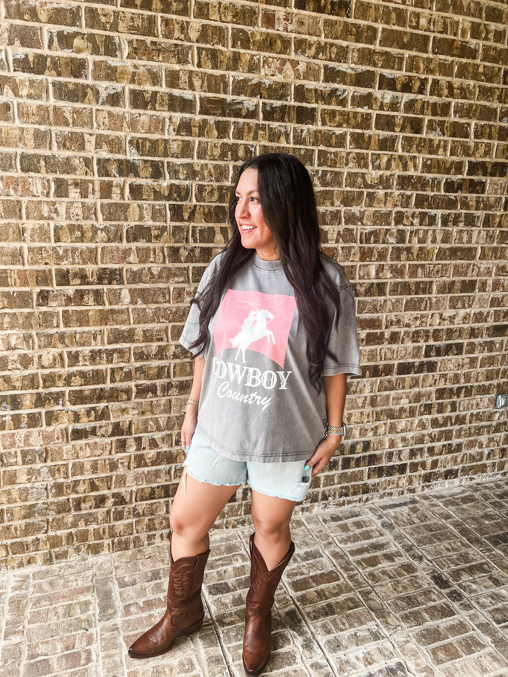 A woman stands against a brick wall wearing a gray graphic t-shirt with a pink horse and cowboy print and the words 'COWBOY Country', light denim shorts, and brown cowboy boots.