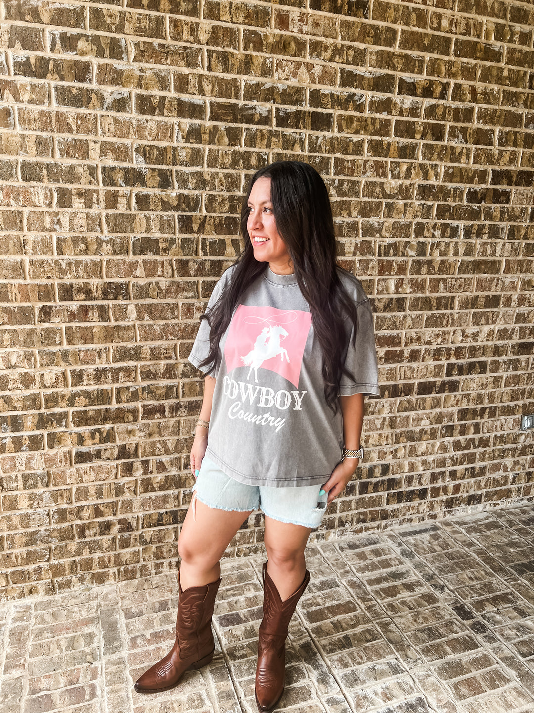 A woman stands against a brick wall wearing a gray graphic t-shirt with a pink horse and cowboy print and the words &