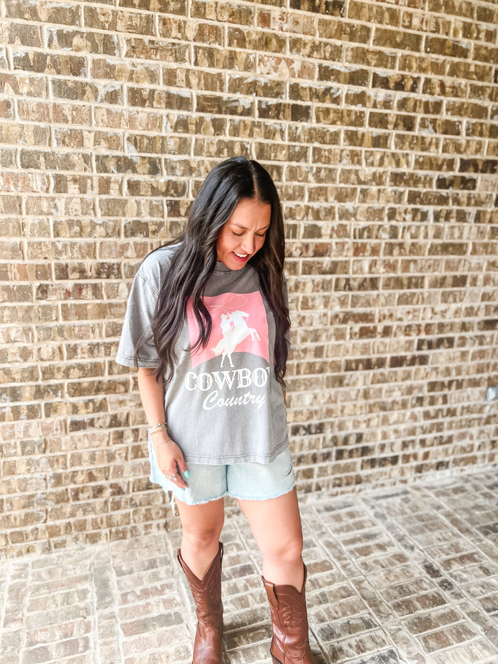 A woman wearing a gray graphic t-shirt with a pink and white cowgirl design, paired with light denim shorts and brown leather cowboy boots, standing in front of a brick wall.