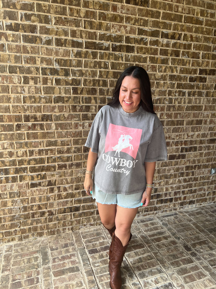 A woman wearing a gray graphic t-shirt with a pink cowboy logo and the words &