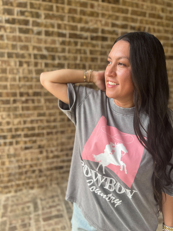 A woman wearing a gray graphic t-shirt with a pink and white cowboy design, posing in front of a brick wall.