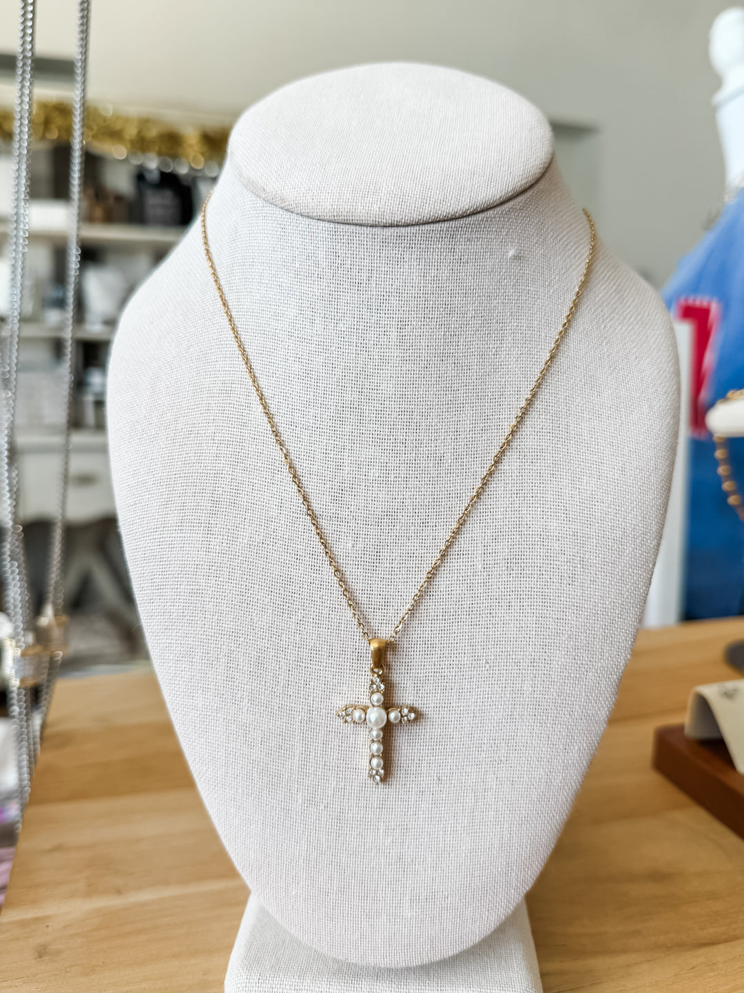 A gold cross pendant necklace with small clear stones, displayed on a white fabric jewelry bust. The necklace is shown against a softly blurred indoor background with a wooden table surface.
