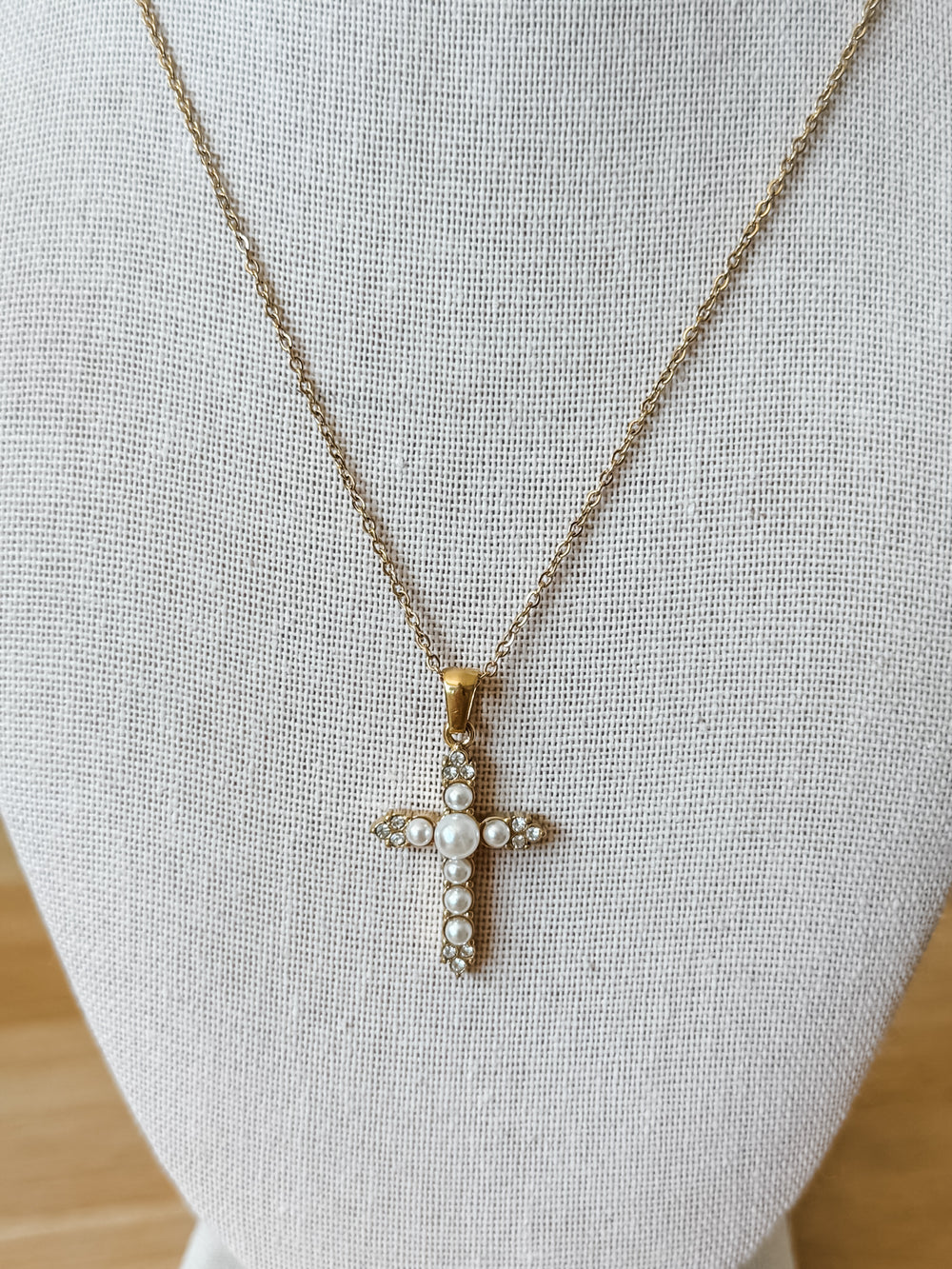 A delicate gold cross pendant necklace with small white pearls, displayed on a textured white fabric jewelry bust.