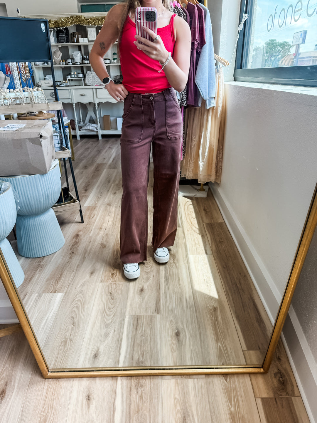 A person taking a mirror selfie in a boutique, wearing a bright red tank top and dark brown wide-leg trousers with white sneakers, set against a background of clothing racks and a wood floor.