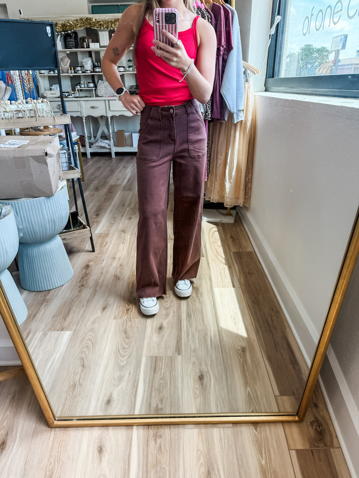 A person taking a mirror selfie in a boutique, wearing a bright red tank top and dark brown wide-leg trousers with white sneakers, set against a background of clothing racks and a wood floor.