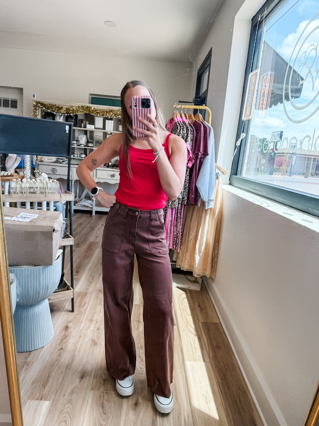 A woman takes a mirror selfie in a boutique, wearing a bright red tank top and high-waisted, wide-leg brown pants with white sneakers.