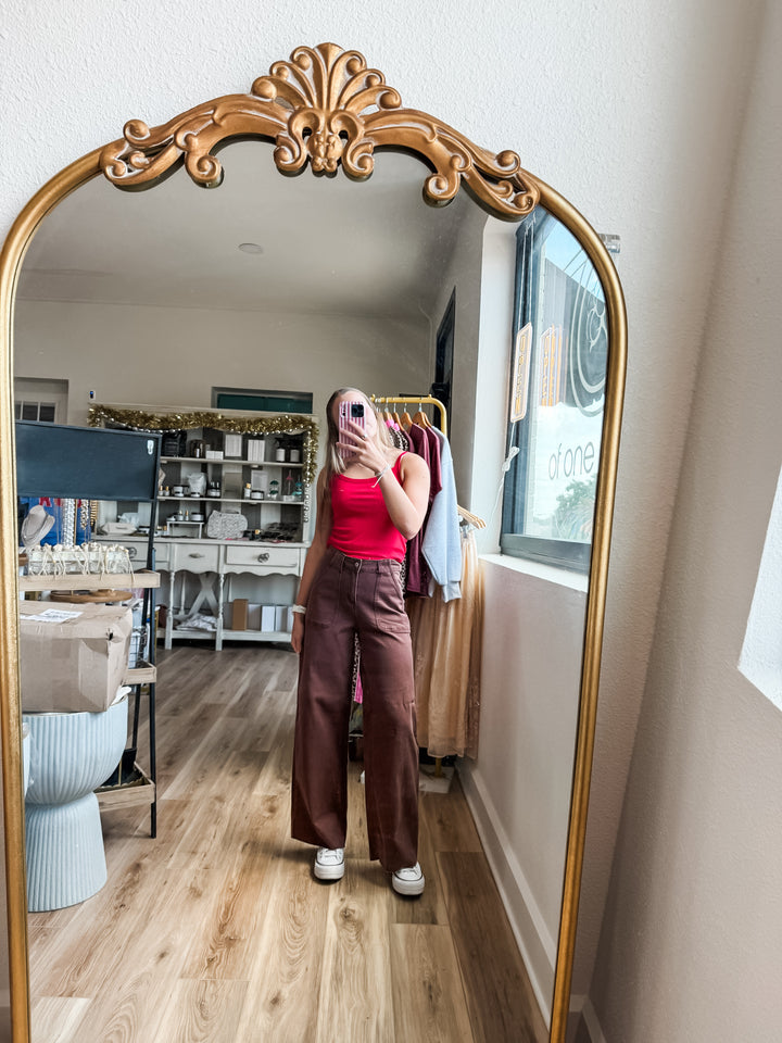 A person taking a mirror selfie in a room, wearing a bright pink tank top and brown wide-leg pants. The photo is taken in front of a large ornate gold-framed floor mirror, showing a closet area and window in the background.