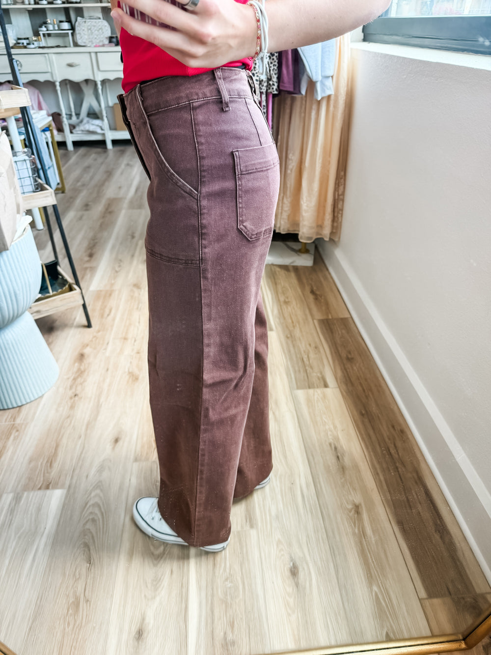 A person standing indoors wearing high-waisted, wide-leg brown pants with a faded wash, paired with a red top and white sneakers. The photo is taken in a mirror showing a home interior with wood floors and furniture in the background.