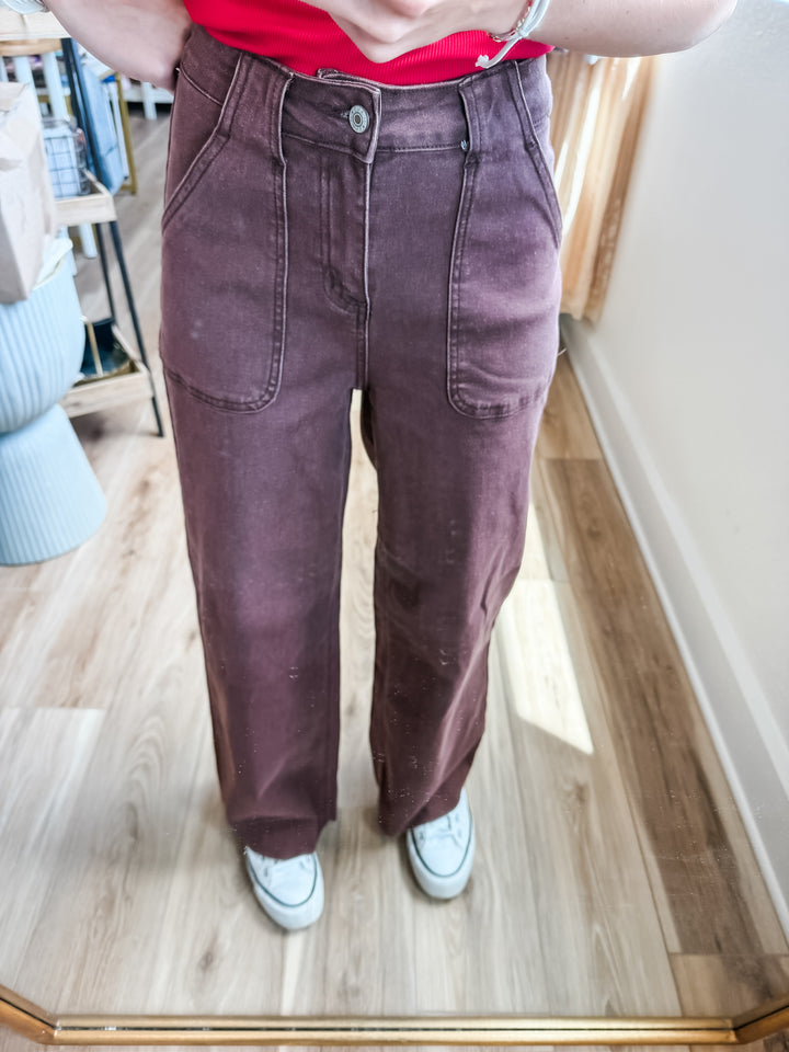 A person wearing a pair of loose, wide-leg corduroy pants in a dusty plum or mauve-brown shade, paired with a red top and white sneakers, standing in a room with a wood floor and mirror.