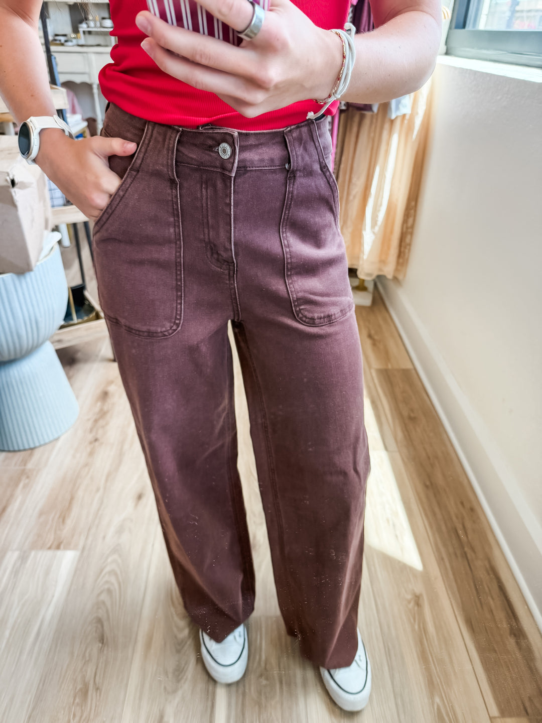 A person wearing a pair of wide-leg, straight-cut pants in a dusty plum or mauve-brown shade, paired with a red top and white sneakers, standing on a light wood floor.