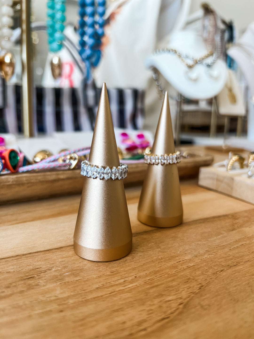 Two gold cone-shaped ring stands displaying a sparkling diamond band ring in gold and silver, set on a wooden tabletop with a softly blurred jewelry store background.