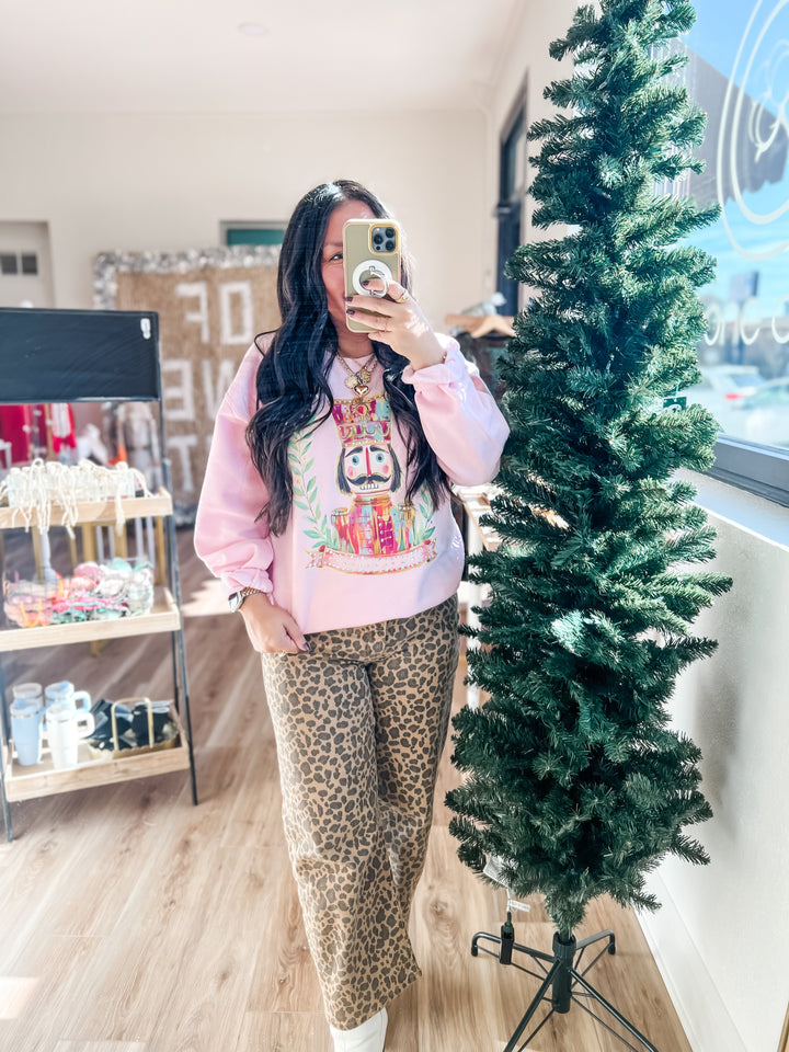 Person wearing a pink sweatshirt with a colorful nutcracker design and leopard print pants standing next to a Christmas tree.