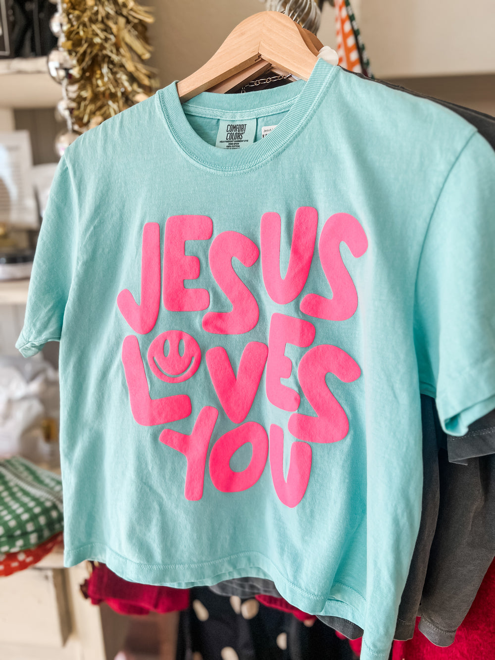 Turquoise t-shirt with 'Jesus Loves You' in pink text on a hanger.