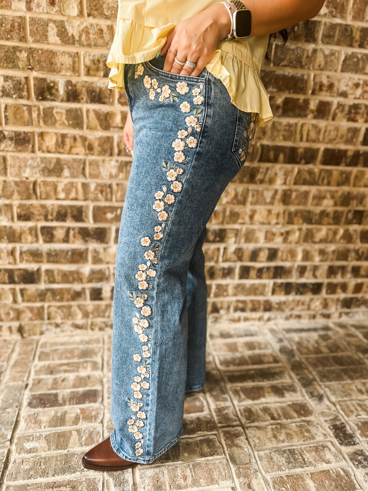 A person wearing blue denim jeans with a vertical floral embroidery design down the left leg, paired with a pale yellow ruffled top and brown boots, standing against a brick wall.