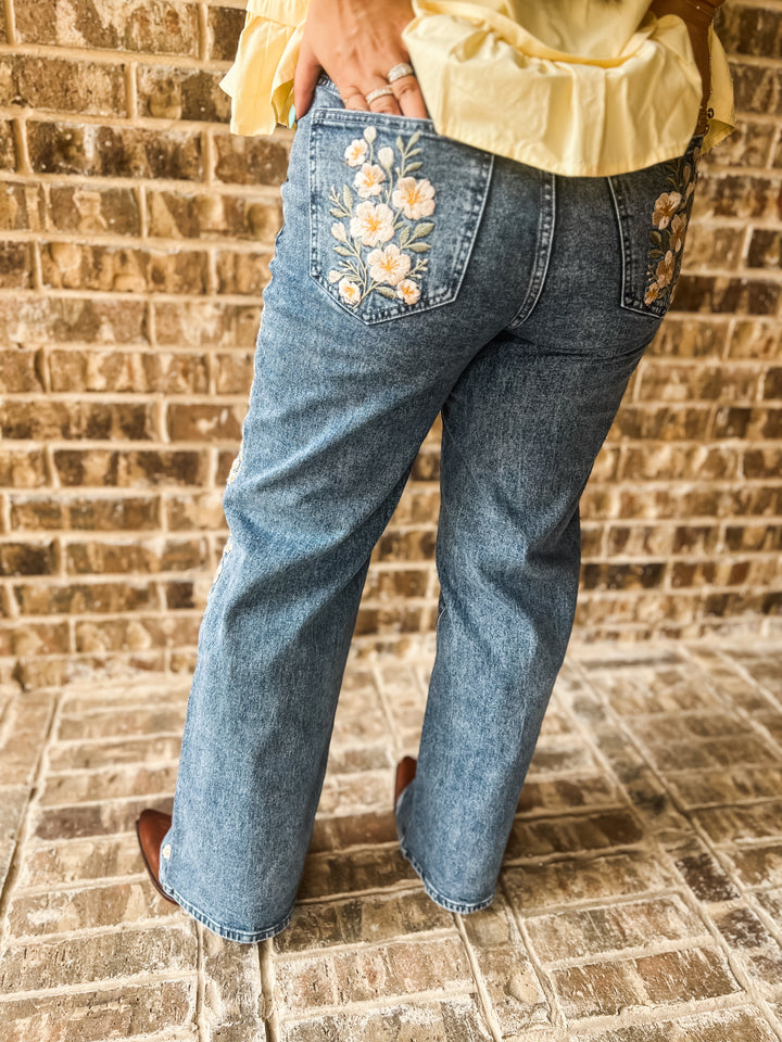 A person wearing blue denim jeans with floral embroidery on the back pockets, standing against a brick wall.
