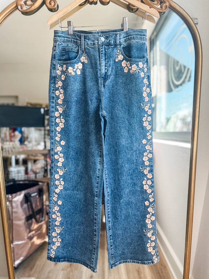 Blue denim jeans with floral embroidery down the outer legs, hanging on a wooden hanger in front of a mirror.