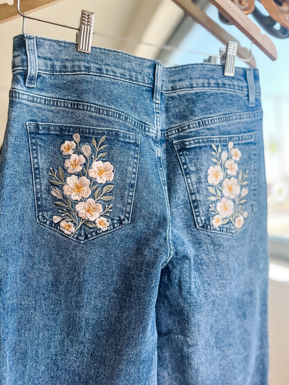 Close-up of the back of blue denim jeans hanging from clips, showing floral embroidery on both back pockets with white, peach, and green flowers.
