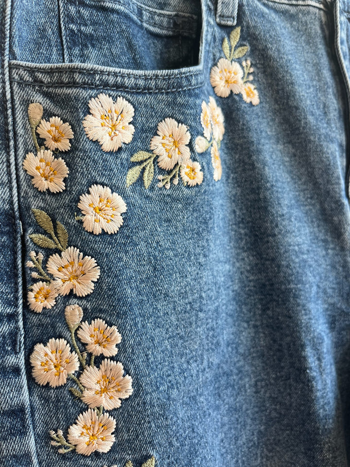 Close-up of a blue denim jeans featuring embroidered cream and yellow flowers with green leaves along the front pocket seam.