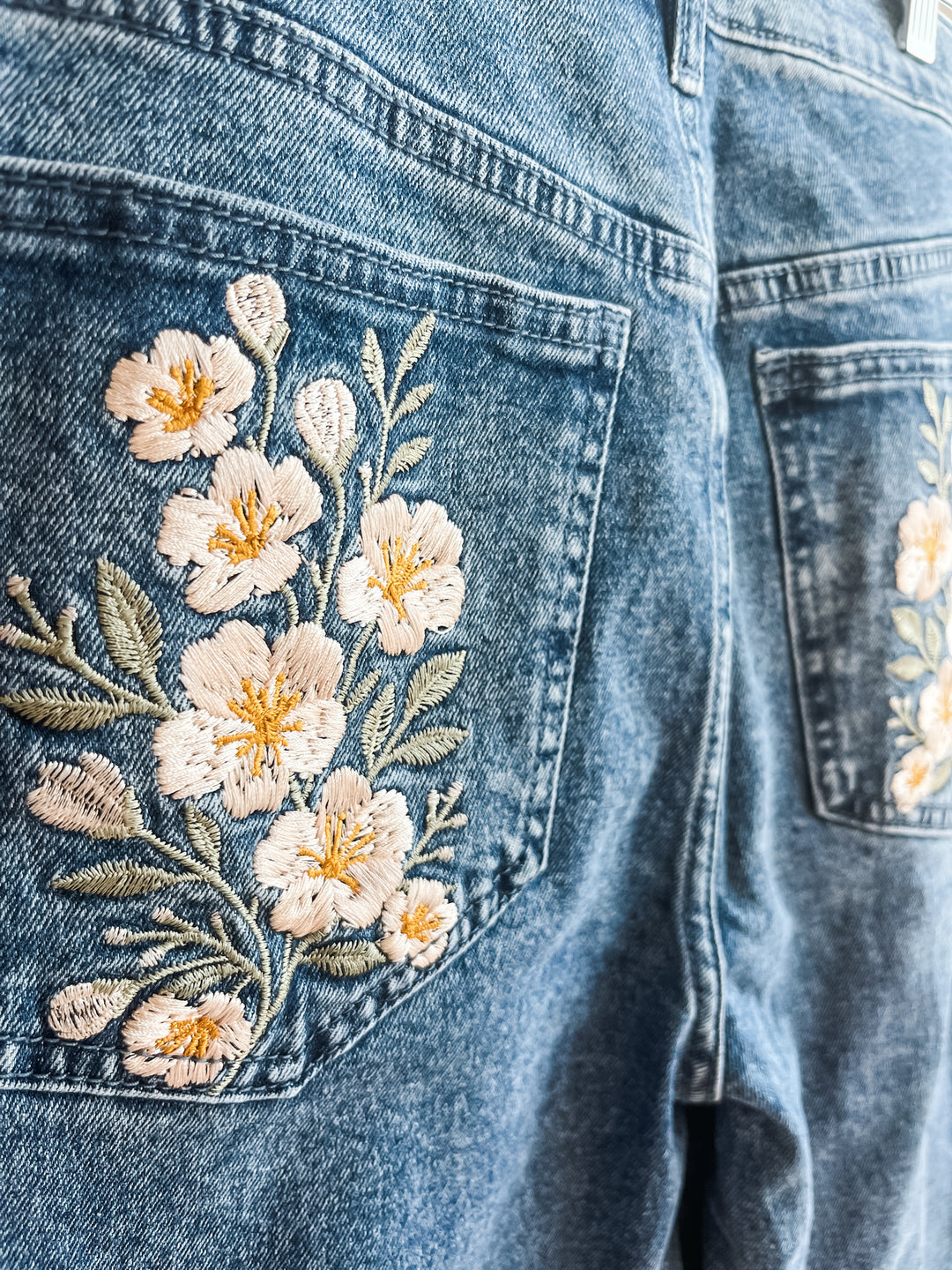Close-up of blue denim jeans showing the back pocket with detailed floral embroidery in cream, pink, and green, alongside another matching embroidered pocket on the right side.