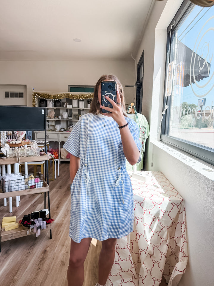 A woman takes a mirror selfie in a boutique wearing a light blue and white gingham short-sleeve dress with a drawstring waist. The shop interior is visible, featuring wooden floors, display tables, and a window.