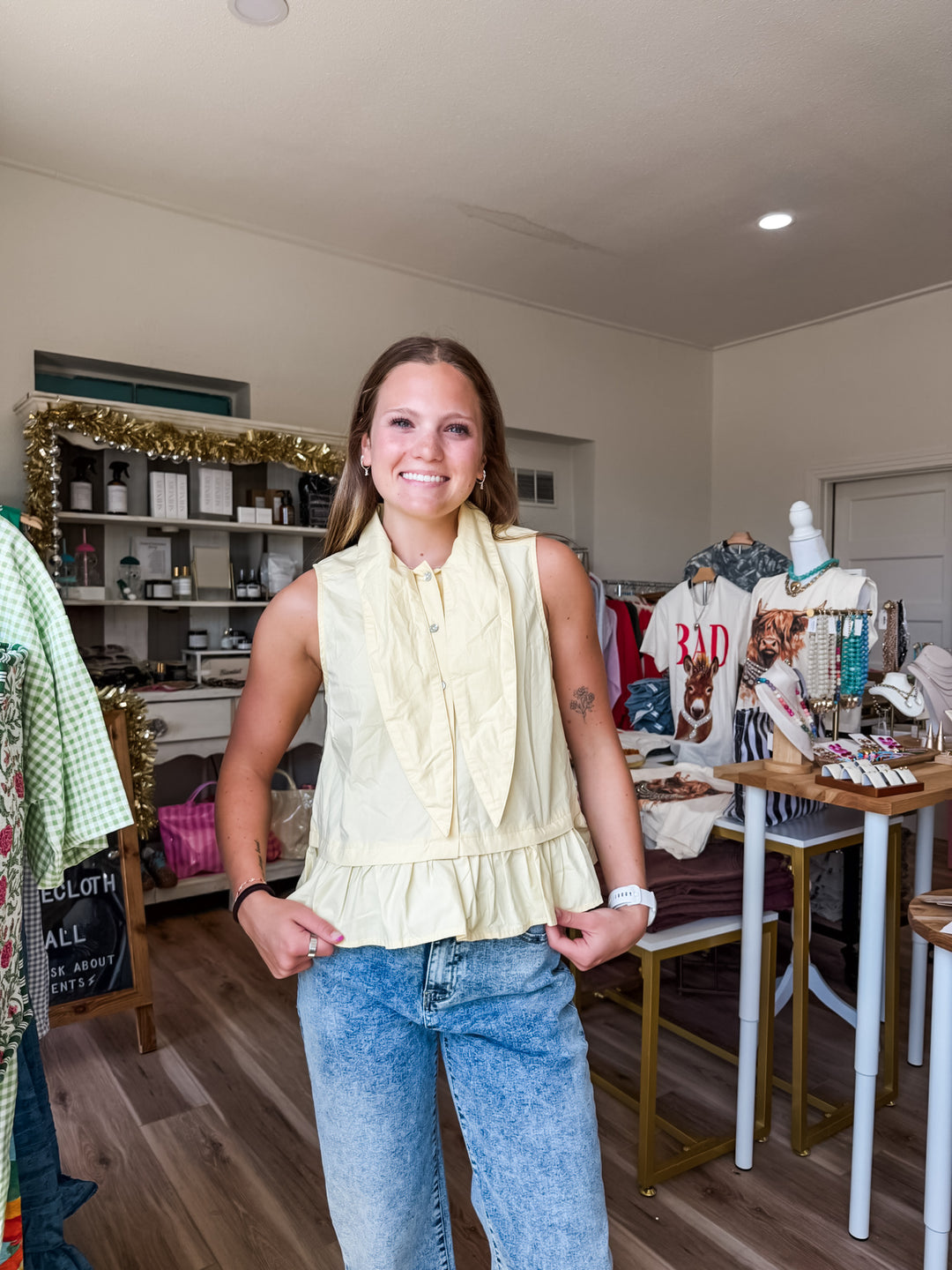 A woman stands in a boutique wearing a pale yellow sleeveless ruffled blouse tucked into light-wash blue jeans. The shop interior has clothing racks, shelves, and a wooden floor.