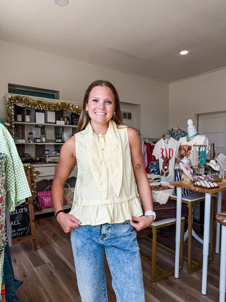 A woman stands in a boutique wearing a pale yellow sleeveless ruffled blouse tucked into light-wash blue jeans. The shop interior has clothing racks, shelves, and a wooden floor.
