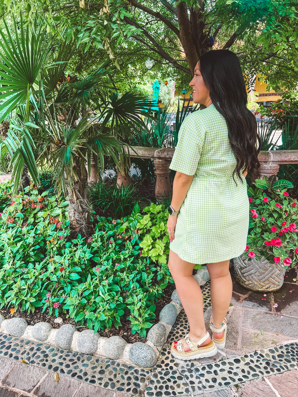 A woman wearing a light green and white gingham mini dress and beige sandals stands in a lush garden setting with tropical plants and flowers.