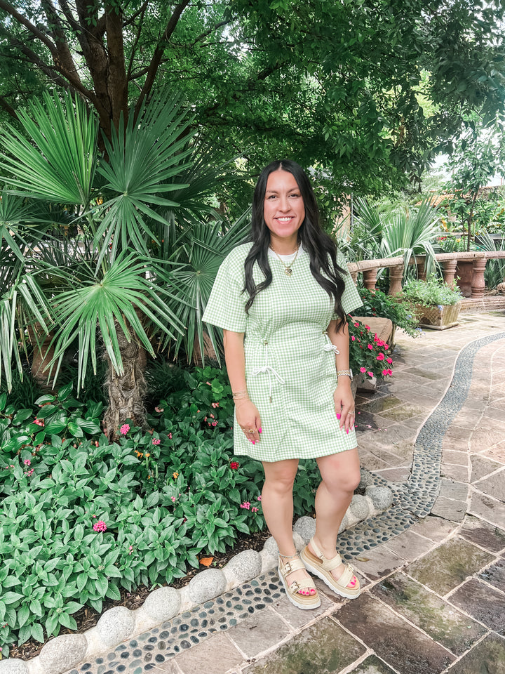 A woman stands on a stone pathway in a lush garden, wearing a light green and white gingham short-sleeve dress with a drawstring waist and beige sandals.