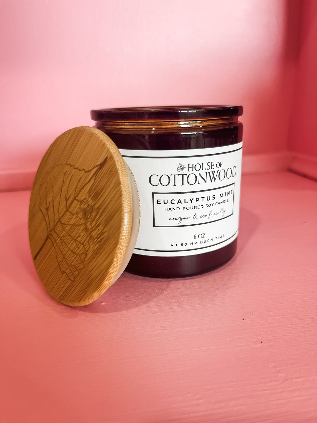 A House of Cottonwood Eucalyptus Mint soy candle in a dark amber glass jar with a white label, shown next to its oval-shaped light wood lid on a pink background.