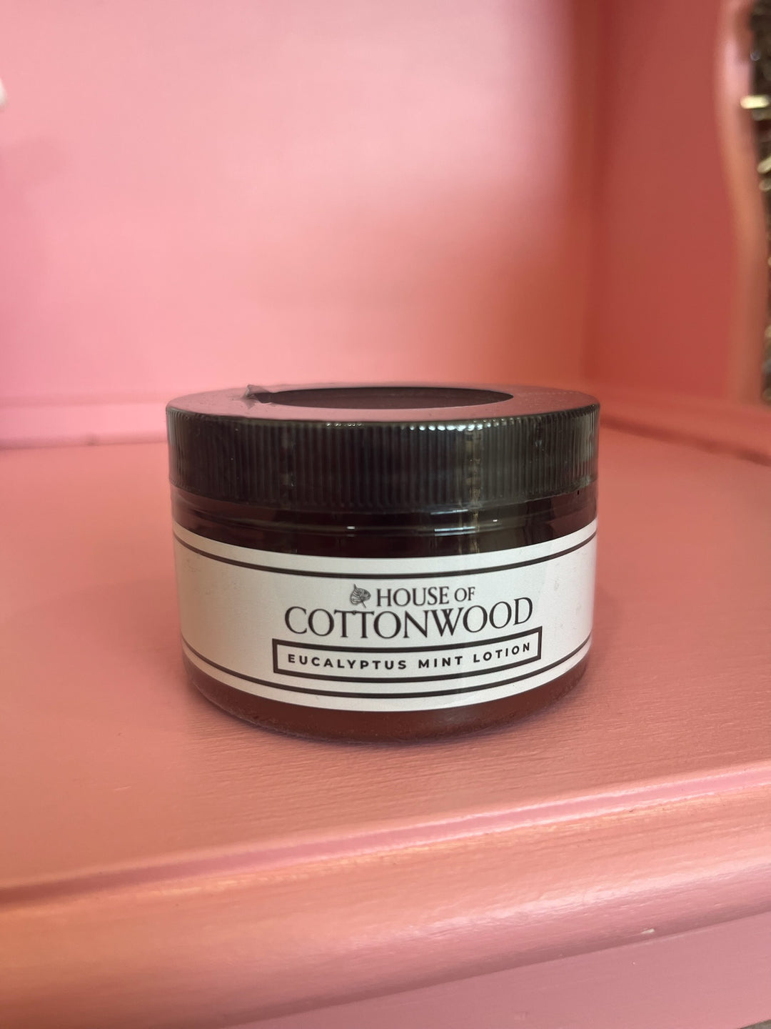 A small jar of House of Cottonwood Eucalyptus Mint Lotion with a white label and black lid, sitting on a pink painted wooden surface.