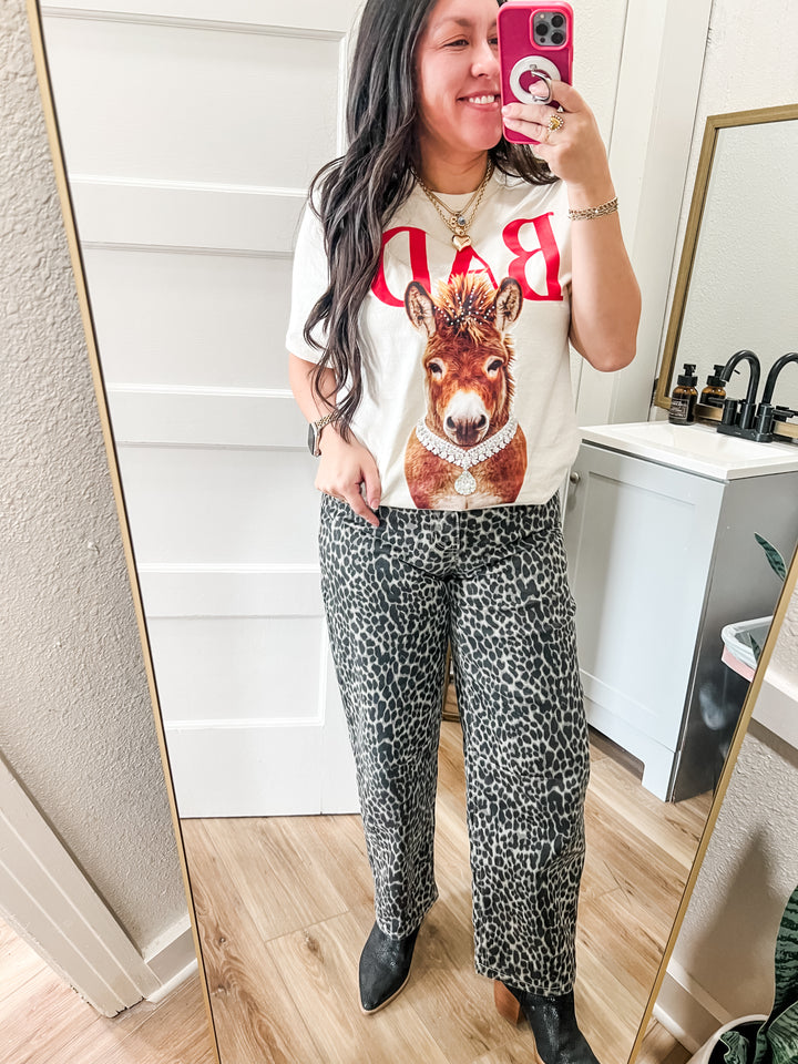 Woman taking a mirror selfie wearing a graphic t-shirt and leopard print pants in a bathroom.