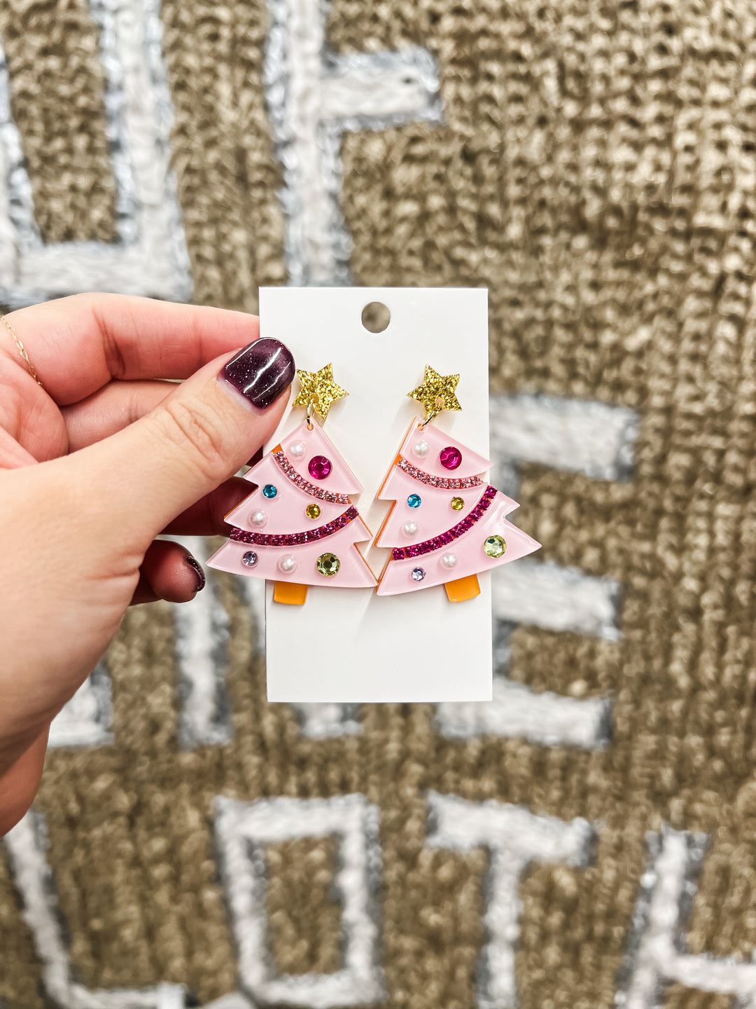Hand holding a pair of pink tree-shaped earrings on a white card against a textured brown background.