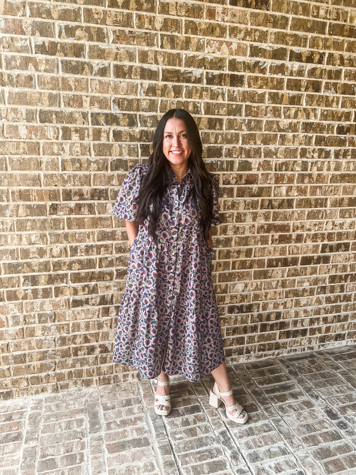A woman stands in front of a brick wall wearing a short-sleeve midi dress with a blue, purple, and pink abstract pattern, paired with white heeled sandals.
