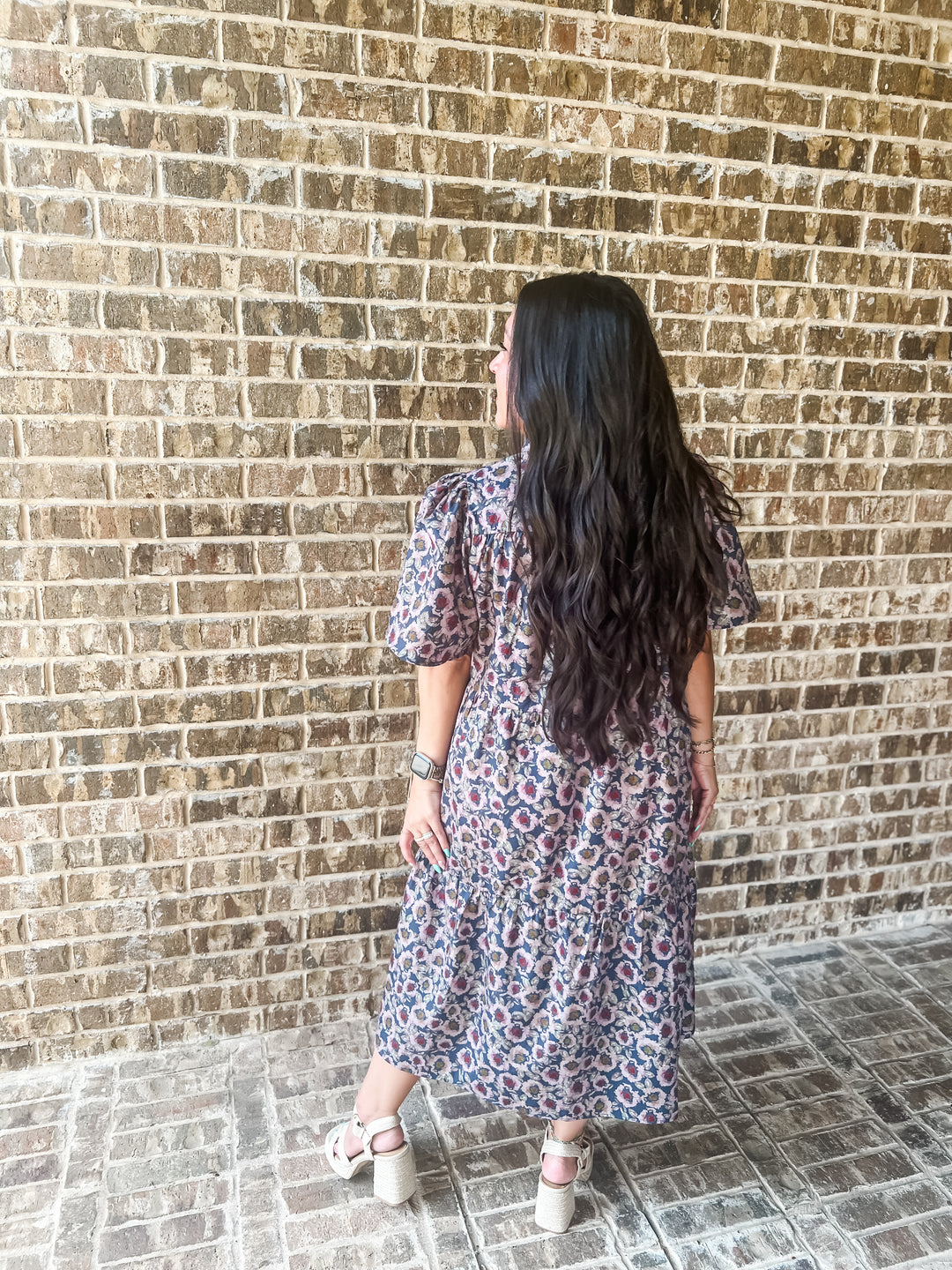 A woman with long, wavy black hair is shown from behind wearing a short-sleeved midi dress with a blue, red, and light pink floral pattern. She is standing on a stone tiled floor against a brown brick wall.
