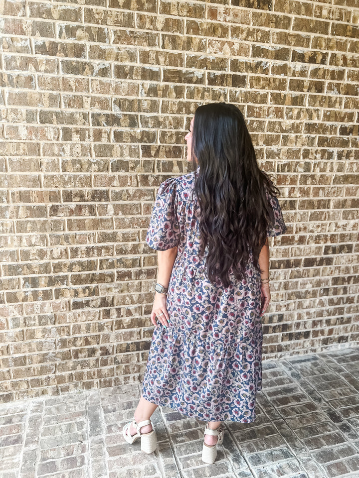 A woman with long, wavy black hair is shown from behind wearing a short-sleeved midi dress with a blue, red, and light pink floral pattern. She is standing on a stone tiled floor against a brown brick wall.