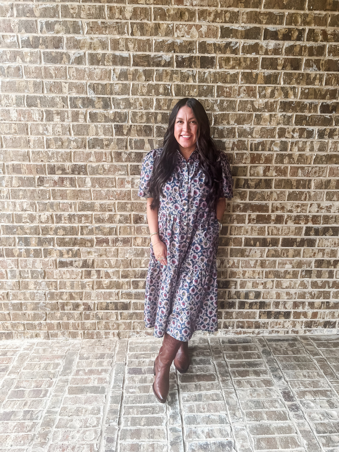A woman stands against a brick wall wearing a short-sleeve cotton button-up dress with a multicolor floral pattern and brown boots.