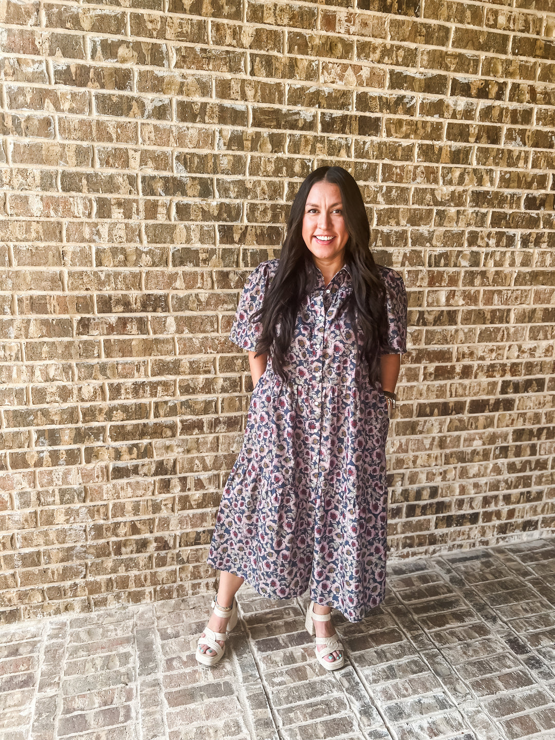 A woman wearing a short-sleeve midi dress with a purple, pink, and blue floral pattern stands in front of a brick wall.
