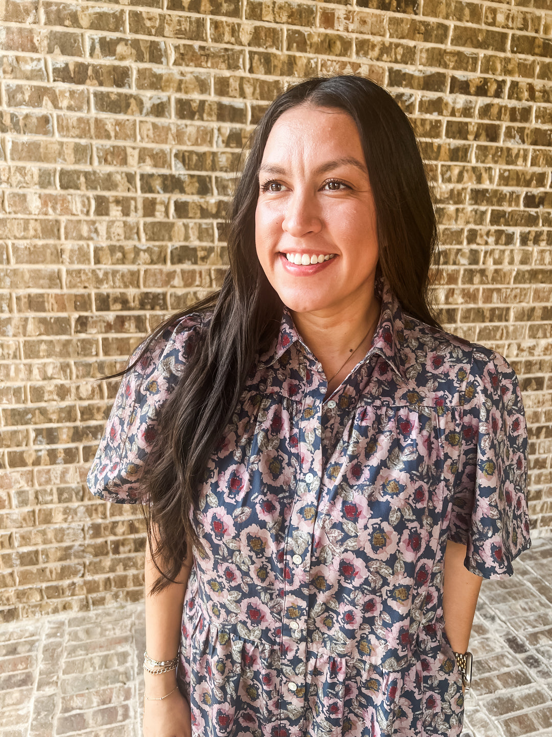A woman with long dark hair smiling in front of a brick wall, wearing a short-sleeve button-up shirt with a blue, pink, and beige floral pattern.