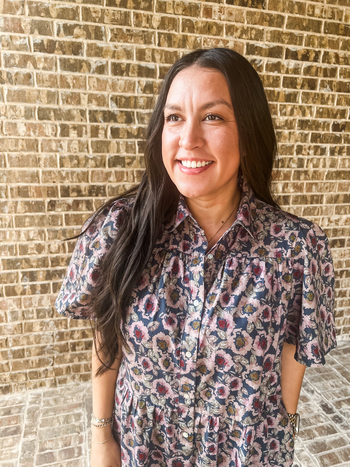A woman with long dark hair smiling in front of a brick wall, wearing a short-sleeve button-up shirt with a blue, pink, and beige floral pattern.
