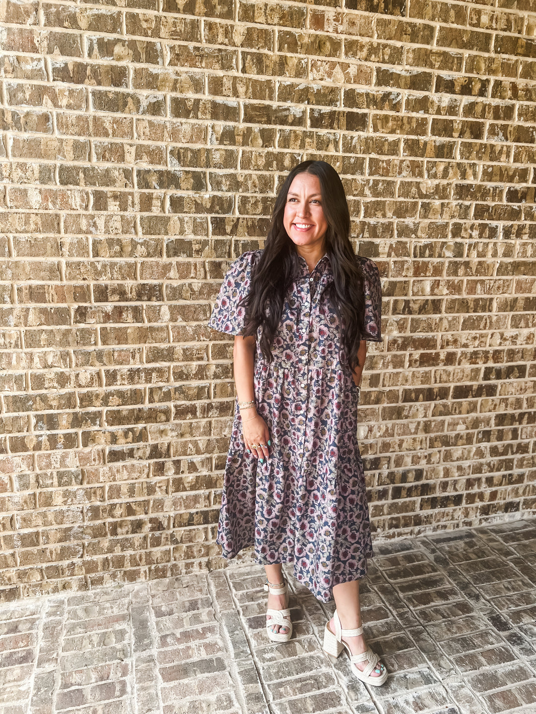 A woman with long dark hair stands in front of a brick wall wearing a short-sleeve midi dress with a purple, pink, and blue floral pattern, paired with white heeled sandals.