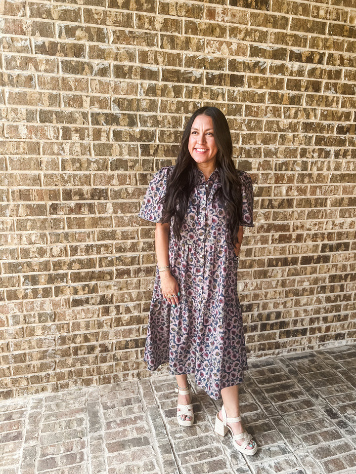 A woman with long dark hair stands in front of a brick wall wearing a short-sleeve midi dress with a purple, pink, and blue floral pattern, paired with white heeled sandals.