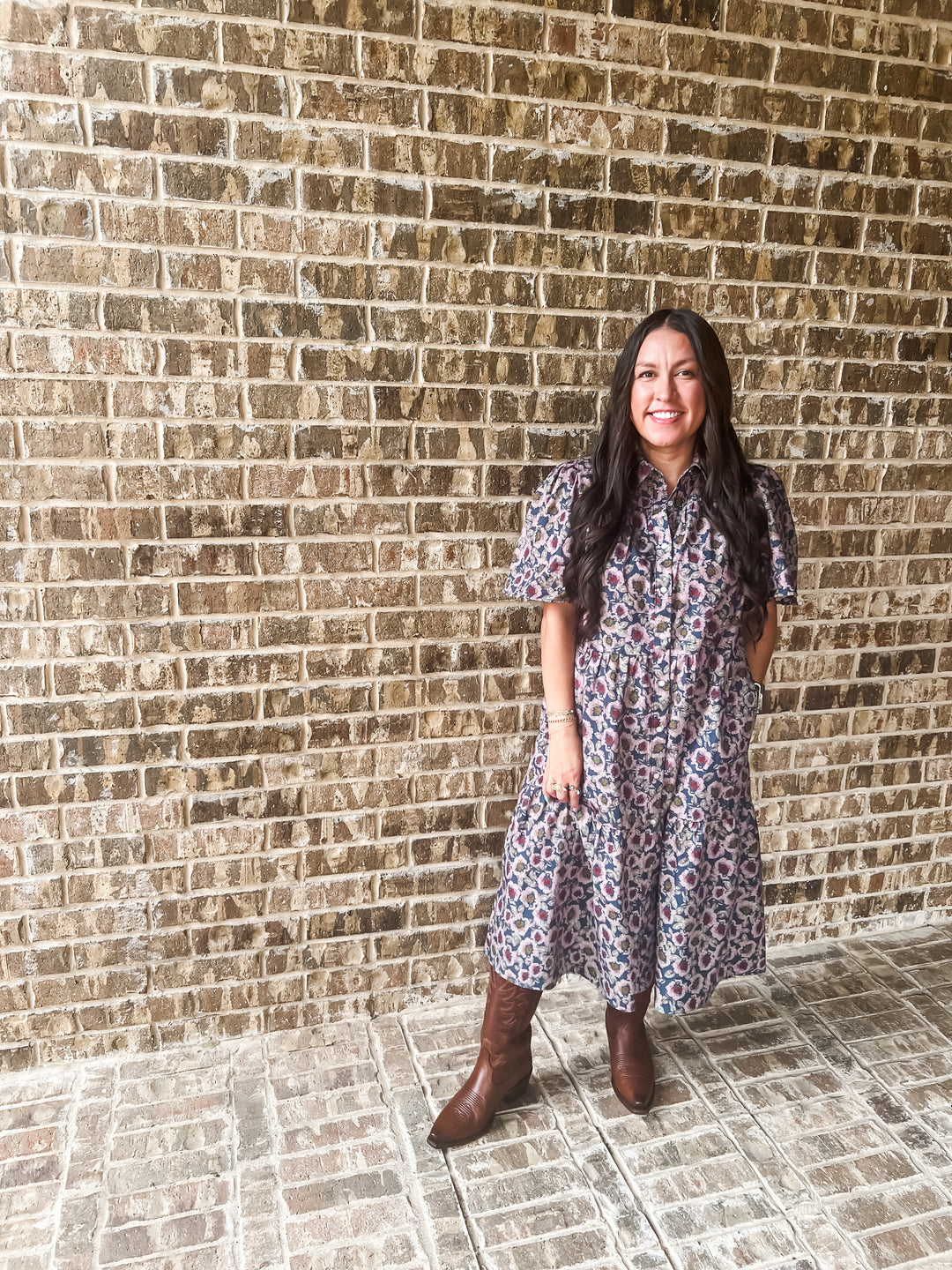 A woman stands in front of a brick wall wearing a short-sleeve midi dress with a blue, red, and white floral pattern, paired with brown leather boots.