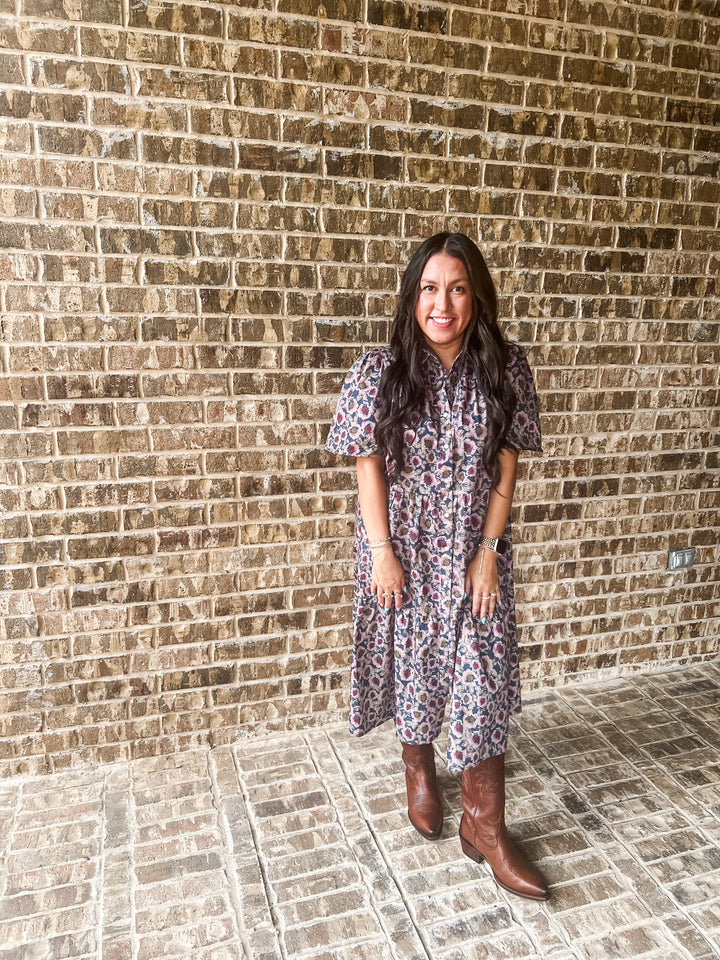 A woman stands in front of a brick wall wearing a short-sleeve button-up dress with a small multicolor floral print and brown cowboy boots.