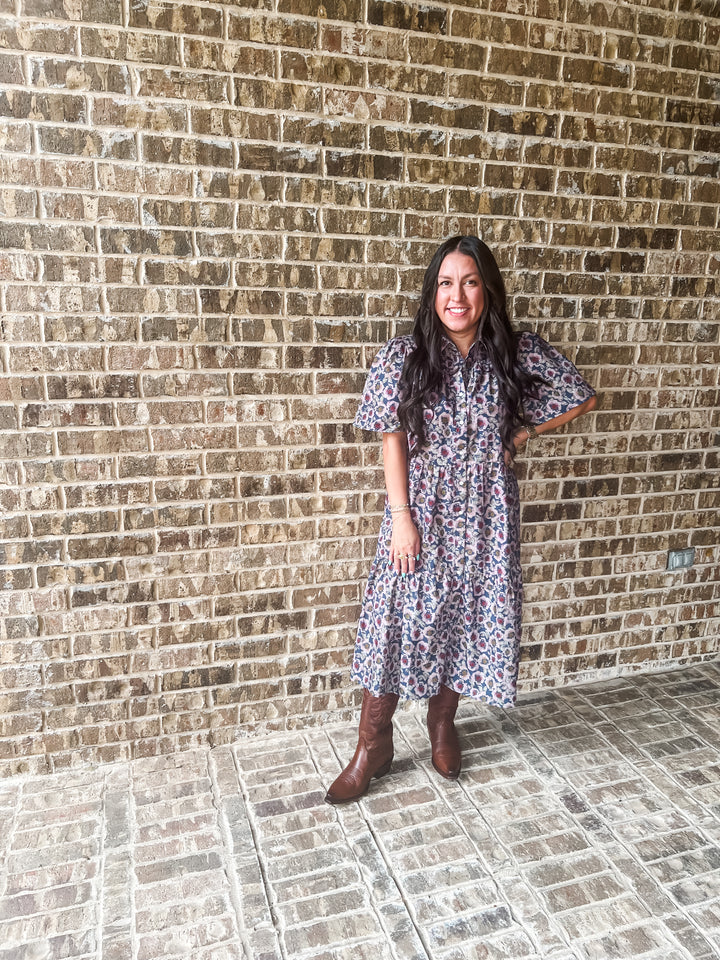 A woman stands in front of a brick wall wearing a short-sleeve, knee-length floral dress with brown boots.