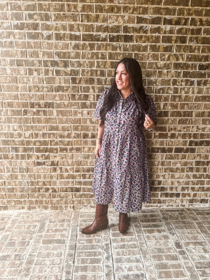 A woman stands against a brick wall wearing a short-sleeve button-front midi dress with a small-scale floral or paisley print in muted purple, pink, and beige tones, paired with brown boots.