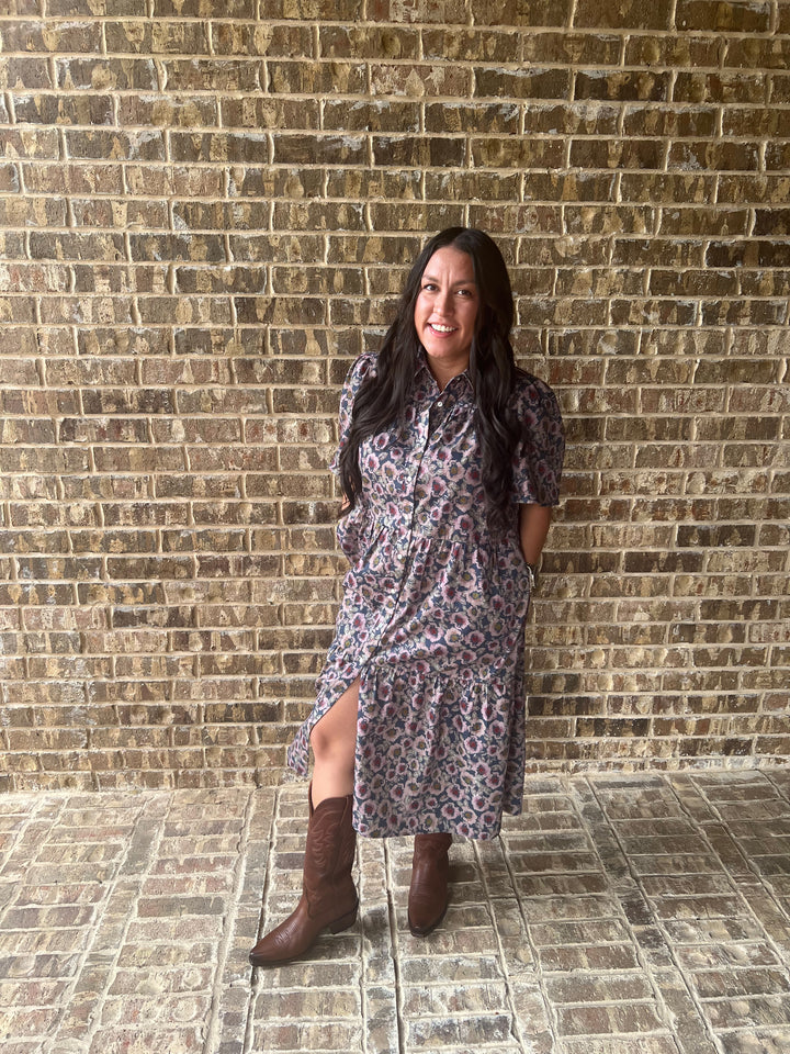 A woman stands against a rustic brick wall wearing a short-sleeve button-up midi dress with a multicolor paisley pattern and brown leather cowboy boots.
