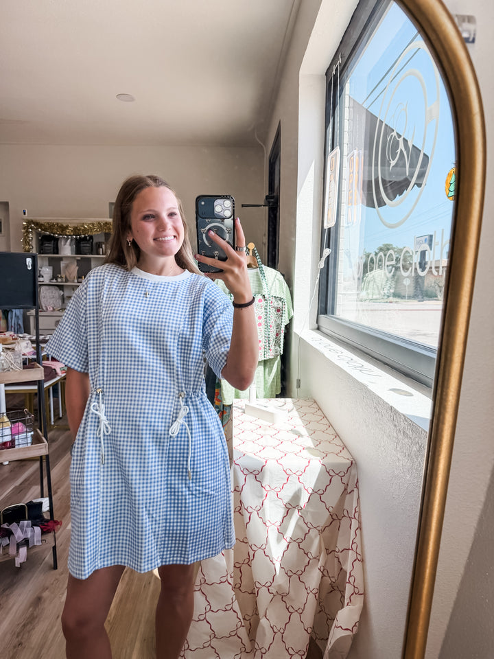 A woman takes a mirror selfie in a boutique while wearing a light blue and white gingham mini dress with a drawstring waist. The shop interior is visible, including a window, clothing rack, and patterned tablecloth.