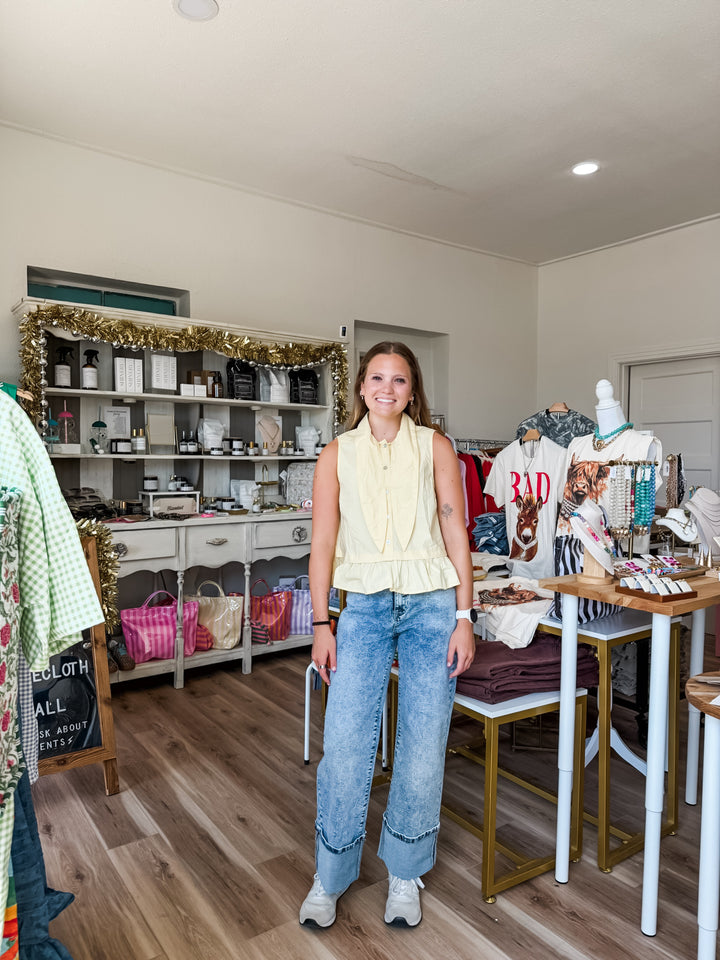 A woman stands in a boutique interior wearing a pale yellow sleeveless ruffled top and light blue wide-leg jeans. Shelves, tables, clothing racks, and store fixtures are visible around her.