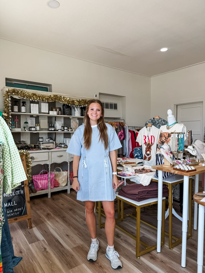 A woman stands in a boutique wearing a light blue and white gingham short-sleeve dress. The shop interior features wooden floors, white walls, shelves with various items, and clothing racks with garments.